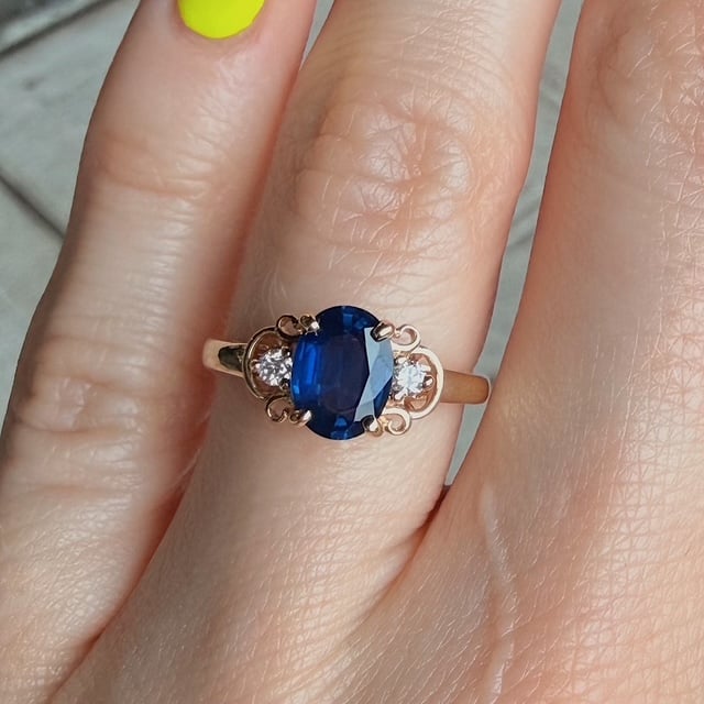 Gold ring with an oval blue sapphire and two small diamonds on a finger with neon-yellow nail polish on the thumbnail side.