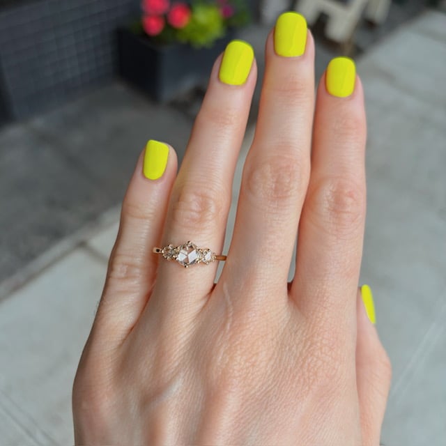 Close-up of a hand with neon yellow nails wearing a gold ring with a central diamond and two side stones on the ring finger.