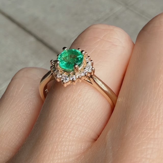 Rose gold ring featuring an oval emerald center stone surrounded by a halo of small diamonds on a finger.