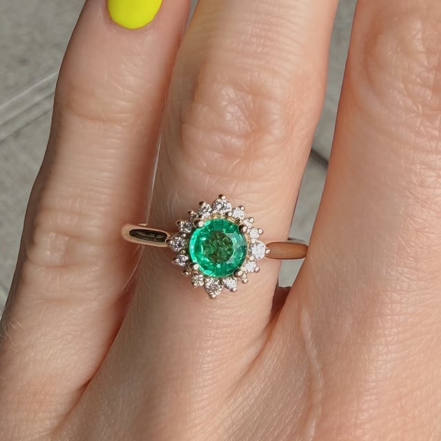Close-up of a rose-gold ring featuring a vivid green central gemstone surrounded by a diamond halo on a finger with neon-yellow nail polish in the background.