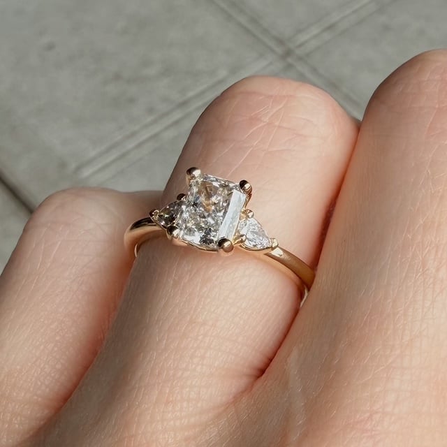 Rose-gold engagement ring with a square-cut center diamond and two side stones on a finger.