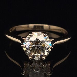 Diamond engagement ring with a round brilliant-cut diamond in a four-prong setting on a slender metal band, against a black background.