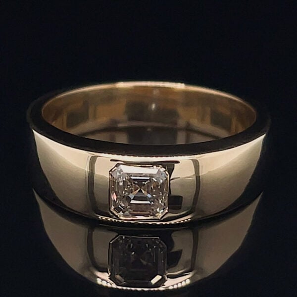 Gold band ring with a bezel-set square-cut diamond, shown on a glossy black surface with its facets reflecting light.