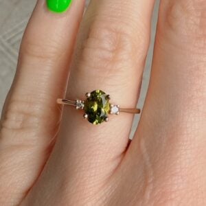 Oval green gemstone engagement ring with a rose gold band and small diamonds on each side, worn on a finger with a bright green nail polish accent.
