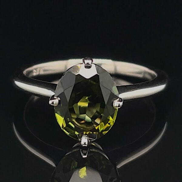 Oval olive-green gemstone ring with a four-prong silver setting, reflected on a black surface.
