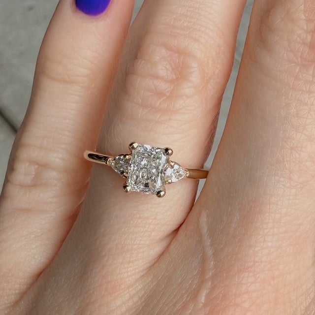 Gold ring with a square-cut center diamond and two side stones, worn on a finger with purple nail polish.