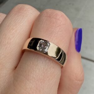 Gold band ring with a square-cut diamond on a finger, close-up shot with a purple-nail foreground