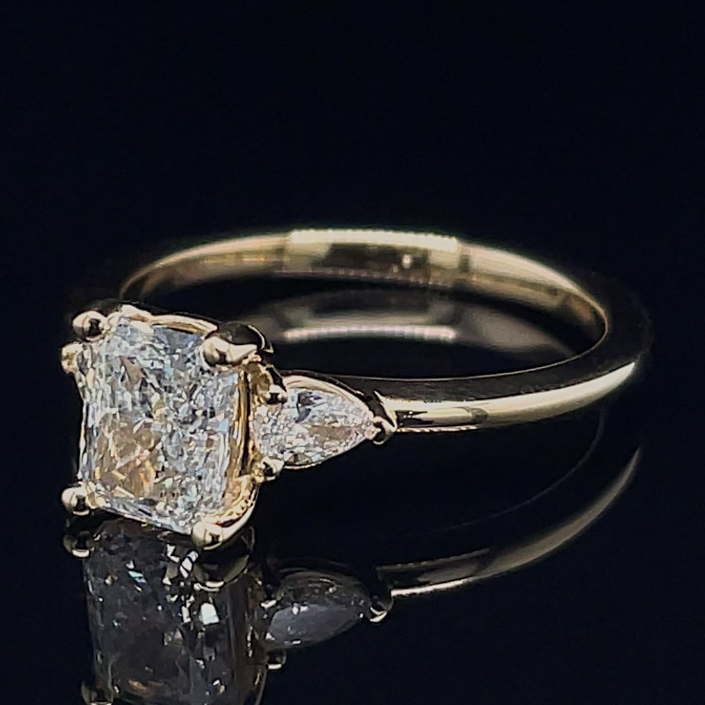 Women’s diamond ring with a cushion-cut center stone and two side stones on a gold band, on a black reflective surface