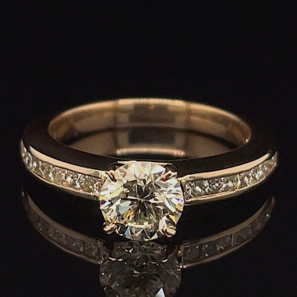 Gold engagement ring with a round center diamond and pavé diamonds along the band, photographed on a dark surface.