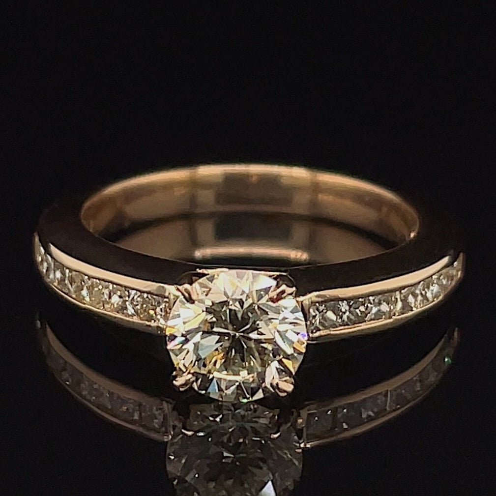Gold engagement ring with a round center diamond and pavé diamonds along the band, photographed on a dark surface.