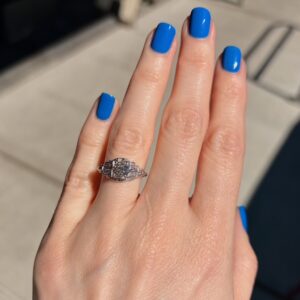 Diamond engagement ring on the ring finger of a hand with bright blue nail polish, photographed in sunlight.