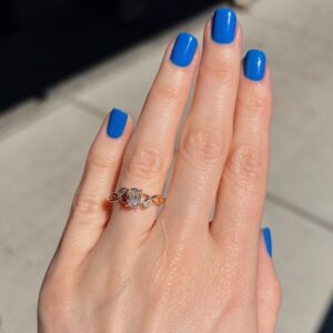 Close-up of a hand with bright cobalt blue nails wearing a gold ring with a central diamond and two side stones on the ring finger.