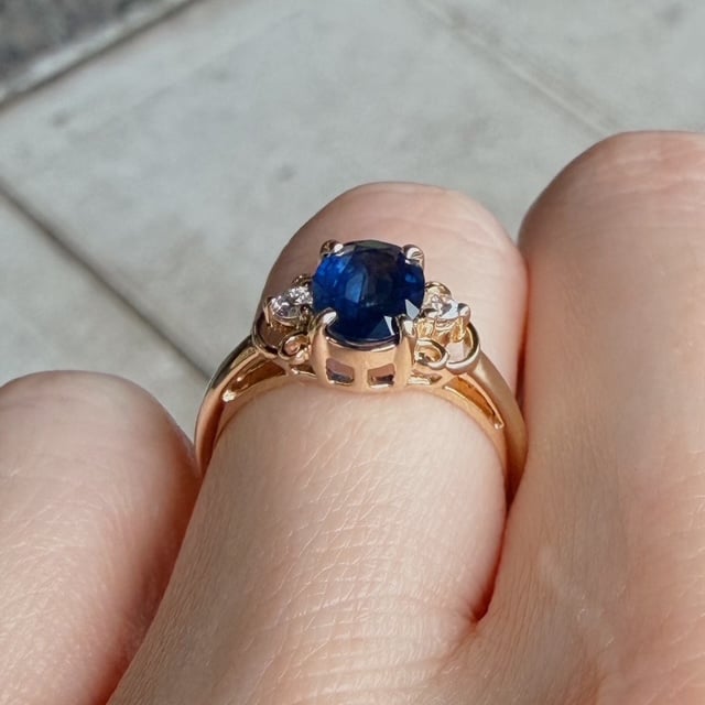 Gold ring with a blue sapphire center stone and two small diamonds on each side, worn on a finger (close-up).