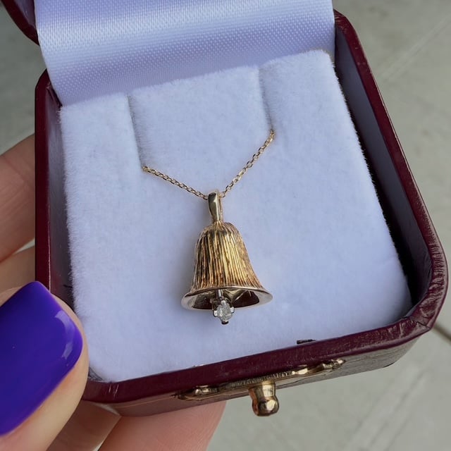 Gold bell pendant necklace with a small gem hanging from its bottom, displayed in a white padded jewelry box.