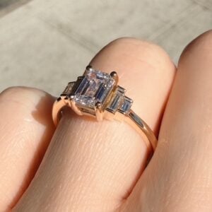 Rose gold ring with a large emerald-cut diamond flanked by baguette diamonds on a finger.