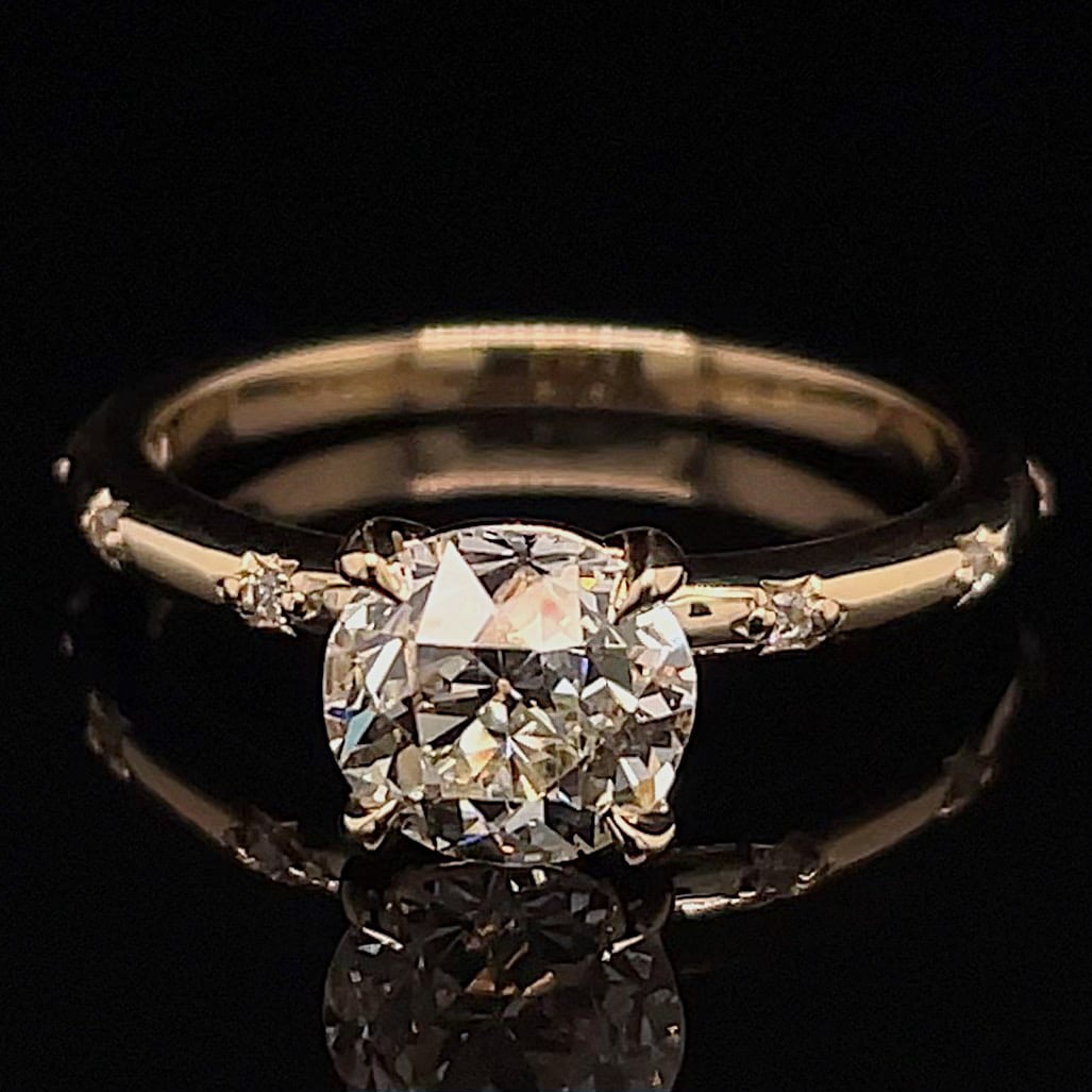 Gold band engagement ring with a large round-cut diamond and small side stones, photographed against a black background, showing sparkle and reflections.
