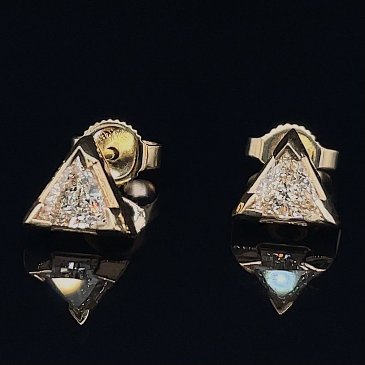 Gold triangle stud earrings with pavé diamonds against a dark background.