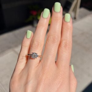Close-up of a hand with mint-green nails wearing a diamond halo engagement ring on the ring finger, outdoors.