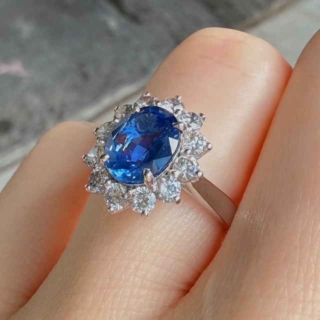 Blue gemstone ring with halo of diamonds on a silver band worn on a finger, close-up detail.
