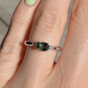 Rose gold ring with an oval green center stone flanked by two small diamonds, worn on a finger with lime-green nail polish.