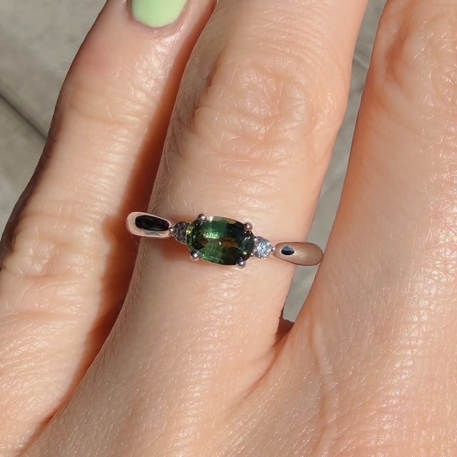 Rose gold ring with an oval green center stone flanked by two small diamonds, worn on a finger with lime-green nail polish.