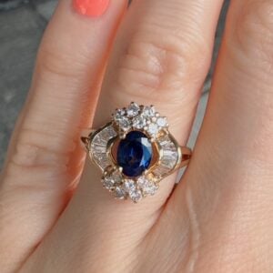 Gold ring with a deep blue oval center sapphire surrounded by white diamonds on a cluster and curved setting.