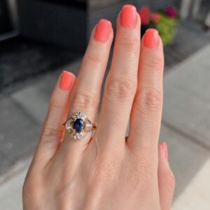Close-up of a hand with coral nail polish wearing a gold ring featuring a blue sapphire centerpiece surrounded by diamonds in a halo setting.