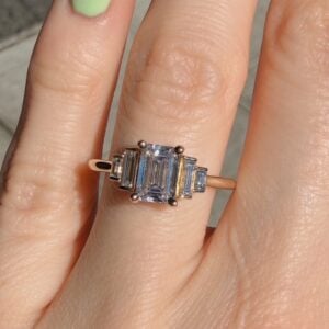 Ring on finger with a central rectangular emerald-cut stone and baguette side stones on a rose-gold band, worn on a manicured finger with green nail polish.