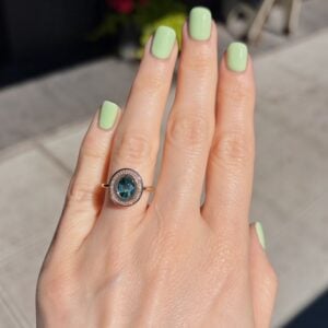 Close-up of a hand with mint-green nails wearing a rose-gold halo ring featuring an oval blue gemstone outdoors.