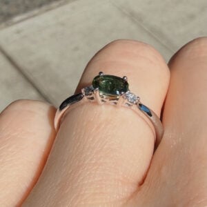 Close-up of a finger wearing a ring set with a dark green oval gemstone and small side diamonds on a metal band.