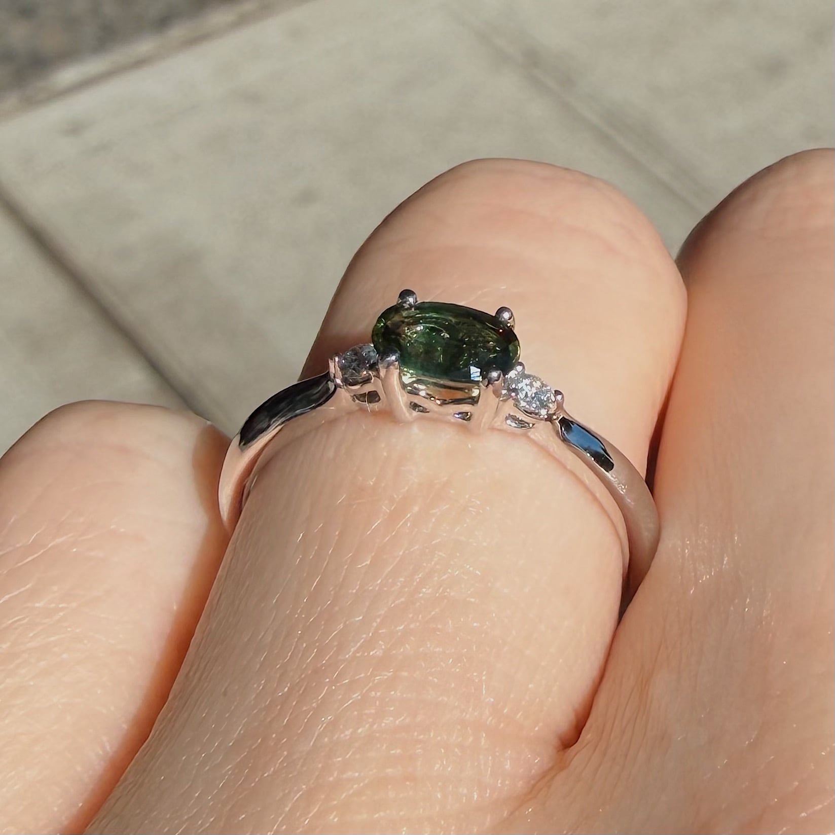Close-up of a finger wearing a ring set with a dark green oval gemstone and small side diamonds on a metal band.