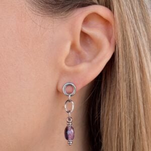 Close-up of a woman's ear wearing a silver dangling earring with two linked hoops and a purple faceted bead, against light skin and hair.