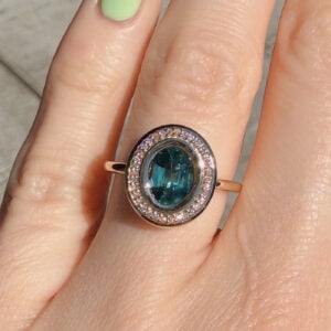 Rose-gold ring with an oval teal gemstone surrounded by a diamond halo on a finger