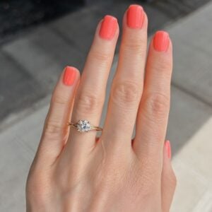 Close-up of a hand with coral nail polish wearing a gold engagement ring with a diamond solitaire on the ring finger.