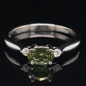 Silver ring with an oval olive-green center stone flanked by two small diamonds on a polished band against a black background.