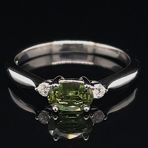 Silver ring with an oval olive-green center stone flanked by two small diamonds on a polished band against a black background.