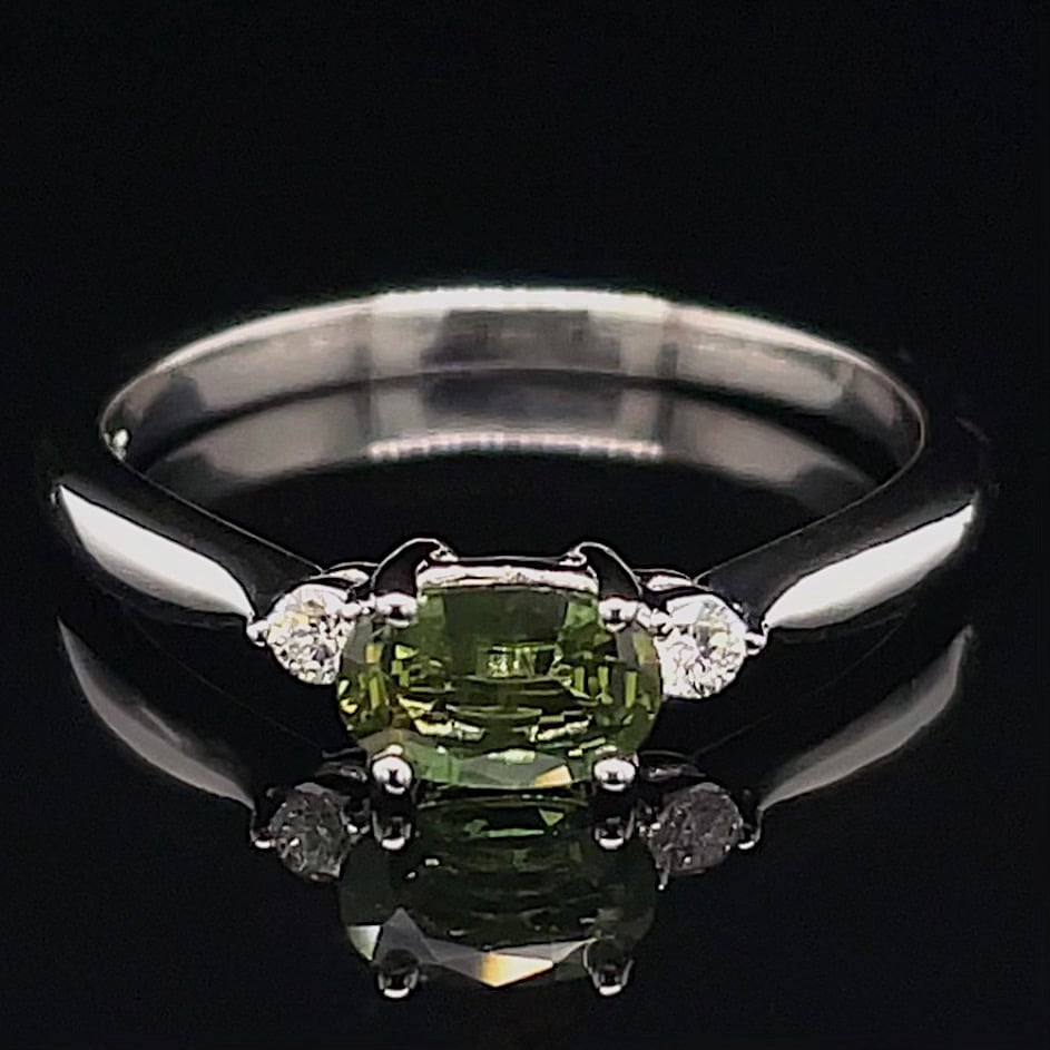 Silver ring with an oval olive-green center stone flanked by two small diamonds on a polished band against a black background.
