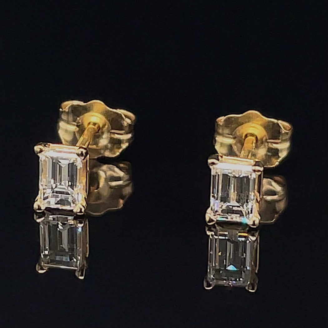 Two gold earrings each with two emerald-cut diamonds stacked vertically, set in prong settings on a dark background.