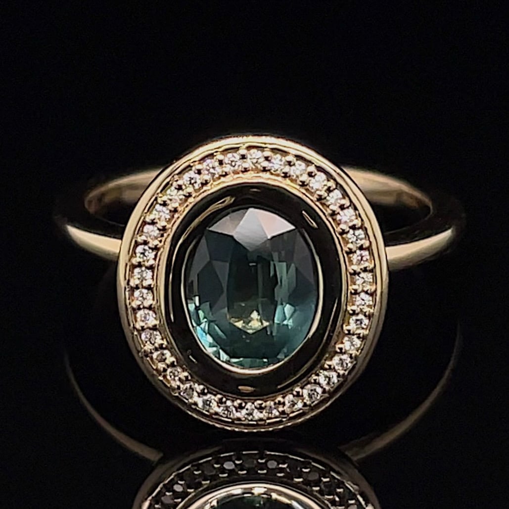 Gold ring with an oval teal center stone, surrounded by a halo of small diamonds on a black background.