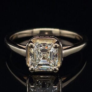 Emerald-cut diamond ring with a four-prong setting on a sleek metal band, photographed against black background.