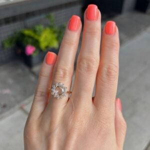 Hand with coral nail polish wearing a halo-style diamond ring on the ring finger, outdoors with a blurred background.