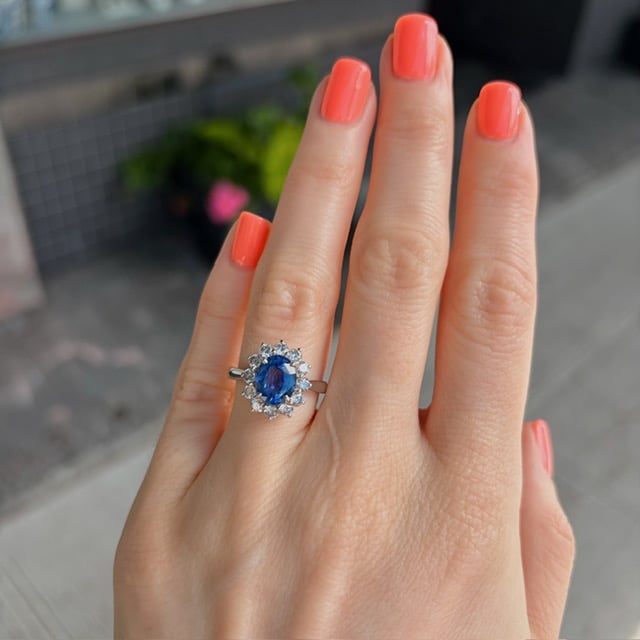 Hand with coral nails wearing a blue gemstone ring surrounded by diamonds on the ring finger.