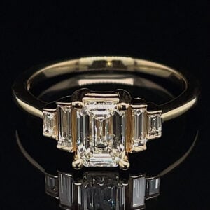 Gold engagement ring with a central emerald-cut diamond flanked by rectangular baguette stones on a black background, close-up.