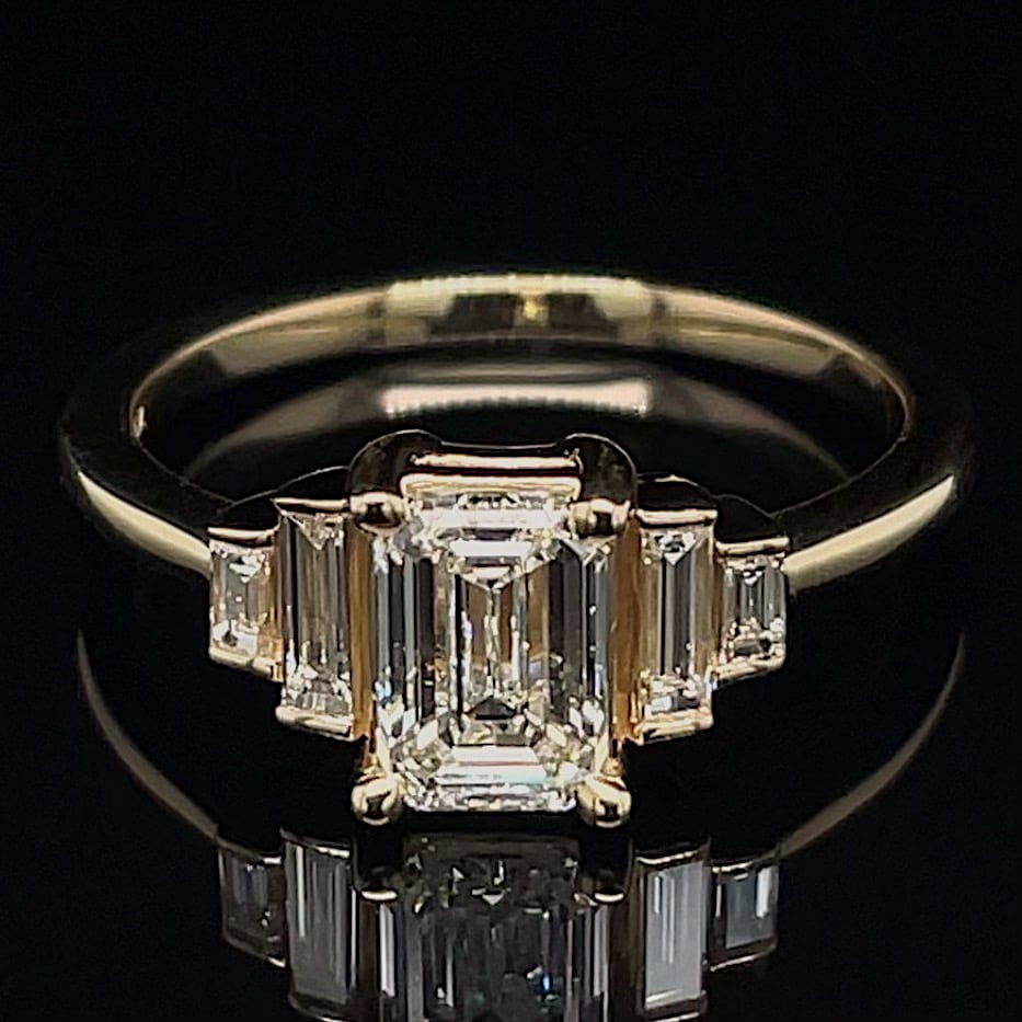 Gold engagement ring with a central emerald-cut diamond flanked by rectangular baguette stones on a black background, close-up.