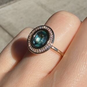 Oval blue-green gemstone ring surrounded by a halo of small diamonds, worn on the finger outdoors.