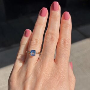 Hand with pink nail polish wearing a gold ring featuring a square blue gemstone in sunlight