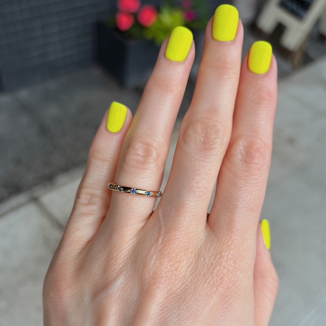 Hand with neon yellow manicure and a thin gold ring with blue stones on the ring finger.