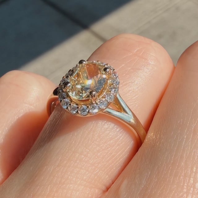 Engagement ring with a halo of small diamonds surrounding a pale yellow oval center stone on a finger in sunlight