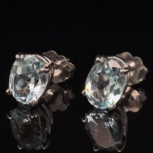 Pair of oval-cut blue gemstone stud earrings in silver prong settings on a black reflective surface.