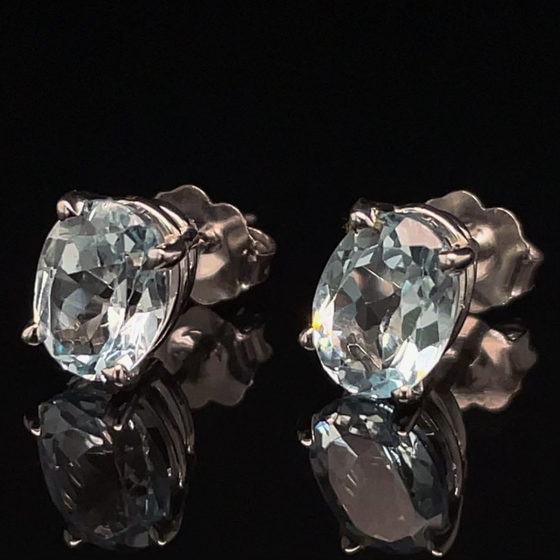Pair of oval-cut blue gemstone stud earrings in silver prong settings on a black reflective surface.